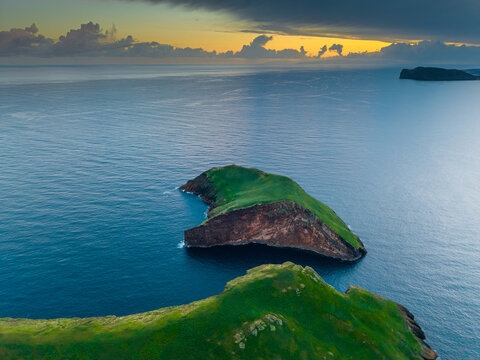 Aerial view of verdant Ilheus das Cabras islands rise from the deep blue Atlantic under a sky brushed with gold and grey, Terceira, Azores, Portugal.