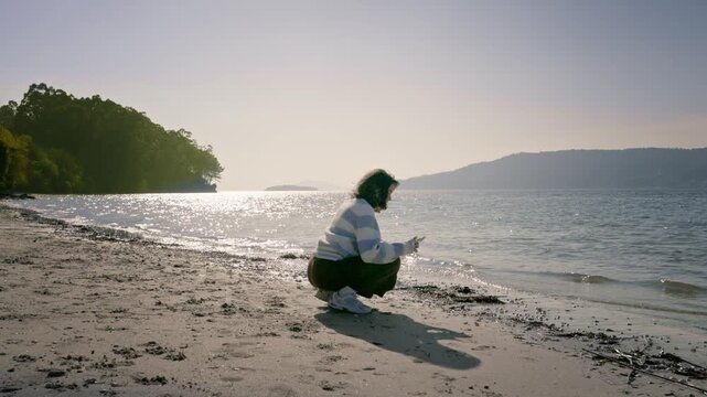 Woman crouching on a sandy shore during a bright afternoon, taking photos with mobile across the calm, shimmering water with mountains visible in the hazy distance