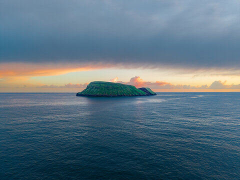 Aerial view of the verdant Ilheus das Cabras islands amidst the vast, dark blue Atlantic ocean under a sky painted with the soft hues of dawn, Terceira, Azores, Portugal.