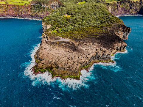 Aerial view of the rugged Ilheus das Cabras islet where dark volcanic rock meets the crashing turquoise waves, Terceira, Azores, Portugal.