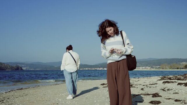 Two female friends spending leisure time together on a sandy shoreline, one looking at the water while the other inspects a seashell against a clear blue sky and a gentle breeze