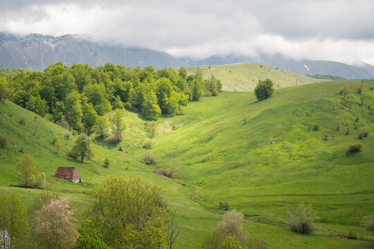 View of verdant hillsides meet a quaint, red-roofed house nestled amidst the vibrant greenery, with distant mountains kissed by clouds, Foca, Bosnia and Herzegovina.