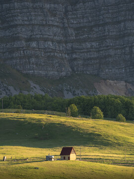 View of a solitary red-roofed house nestled in the green meadow, under the shadow of the rugged, layered cliffs of Zelengora, Foca, Bosnia and Herzegovina.