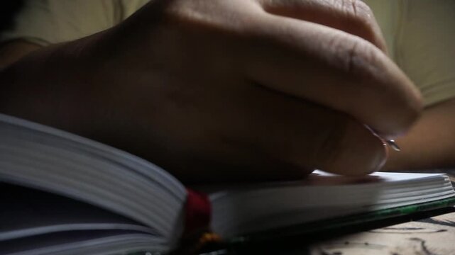 A close up shot shows a male opening a green notebook and preparing to write with a pen in a dimly lit room, creating a moody and focused atmosphere.