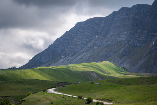 View of a winding road cutting through vibrant green meadows under the shadow of towering, layered mountains, Foca, Bosnia and Herzegovina.