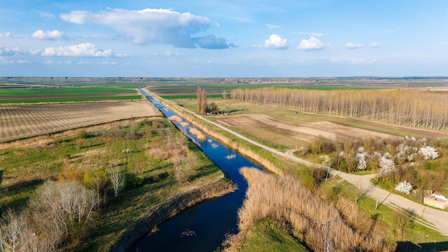 Aerial view of a tranquil canal cutting through the vibrant patchwork of fields and forests under a vast sky, Krcedin, Vojvodina, Serbia.