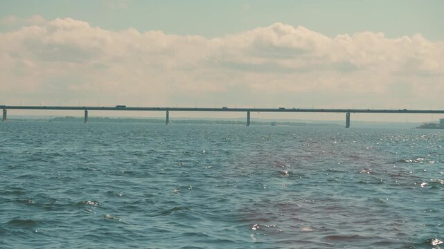 Serene landscape of a long bridge spanning a vast river with gentle waves. Cars travel across the highway under a soft, cloudy sky. Muted, peaceful color palette