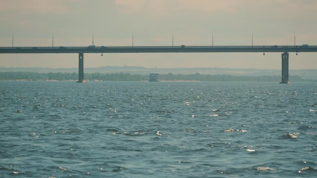 A serene landscape view of a long concrete bridge with cars crossing a vast, shimmering river. The distant shore is shrouded in mist, creating a calm and peaceful mood. Muted colors and soft light