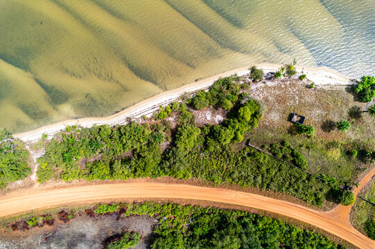 Aerial view of sandy shores kissed by turquoise waters, meeting lush greenery alongside a terracotta-hued road, creating a vibrant contrast., Monrovia, Liberia.