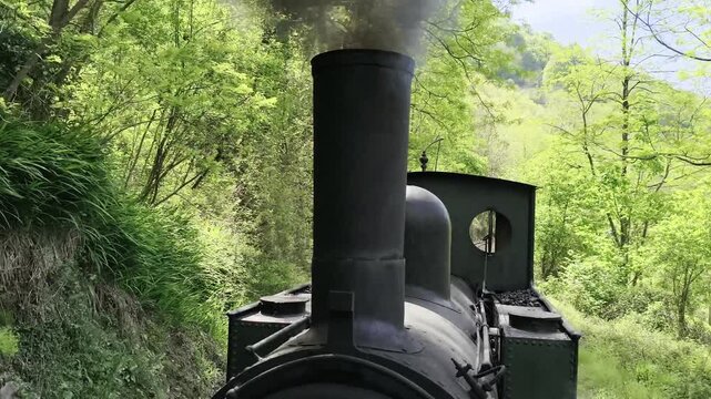 Vista en primer plano de la chimenea de una locomotora de vapor antigua expulsando humo denso mientras circula por una v&iacute;a rural bajo un cielo despejado.