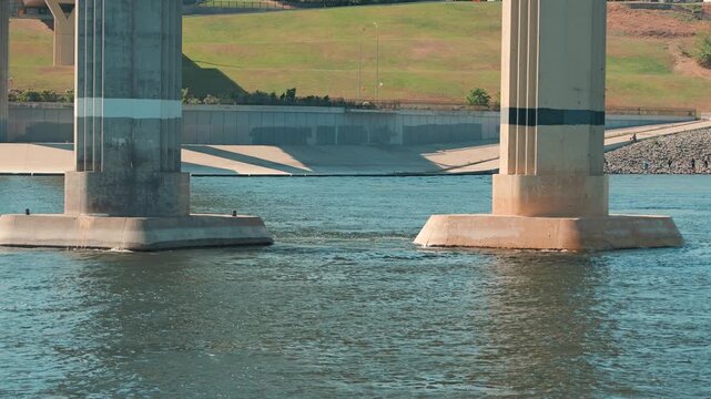 Urban landscape featuring massive concrete bridge pillars in a river. Bright sunlight casts strong shadows on the embankment and green hill. A calm, structural view of infrastructure and nature