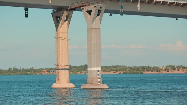 A view from the water looking at massive concrete bridge supports. The calm blue river flows around the piers under a light blue sky. A tranquil scene of modern engineering and transportation
