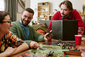 Fototapeta premium Young adult friends playing tabletop role playing game, holding cards and planning moves at home