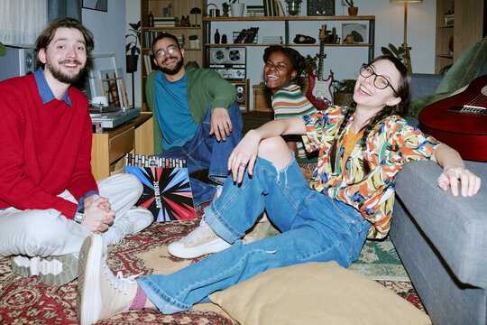 Portrait of diverse young adults sitting on floor, smiling at camera during home gathering with vinyl records and guitars