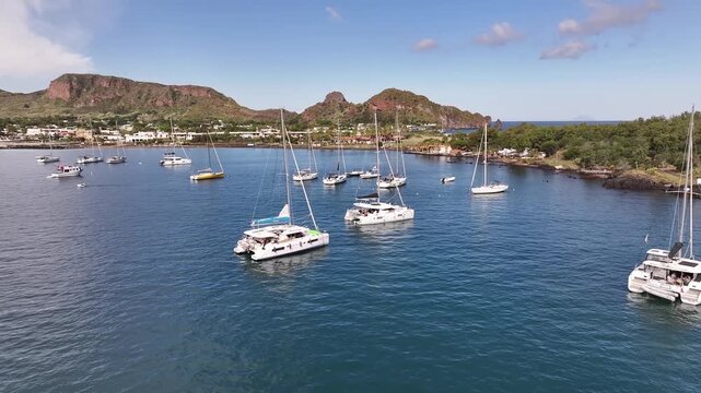 Drone flying backward from anchored yachts revealing scenic bay and Vulcano island coastline in Italy.