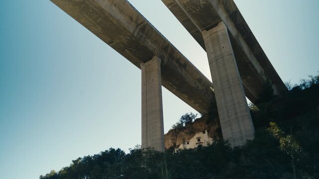 Low angle view of a large concrete viaduct spanning a canyon with towering pillars and a small house beneath. Strong contrast between infrastructure and natural landscape under clear blue sky.