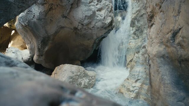 Small waterfall flowing through a rocky canyon, framed by foreground rocks with smooth mineral textures and natural patterns. Cinematic composition with depth, erosion details and flowing water.