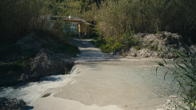Small river flowing over a driveway leading to a rural house surrounded by vegetation. Natural water crossing creating a unique access point in a quiet countryside setting.
