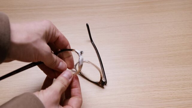 Hand picks up old broken eyeglasses from a wooden desk and removes the lenses from both sides of the frame. DIY repair or recycling process. Real and authentic everyday footage.