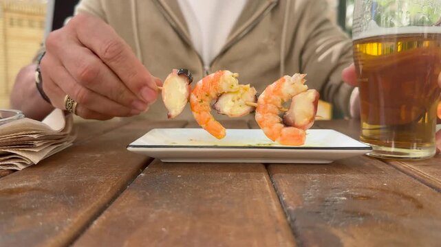 closeup view of the upper body part of a man wearing a hoodie enjoying a spanish traditional skewer with a beer, sitting outdoor on. wooden table,