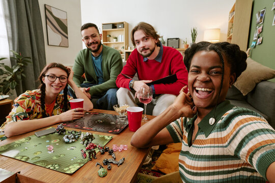 Portrait of diverse young adults playing tabletop game and taking selfie during home gathering, showing friendship and leisure