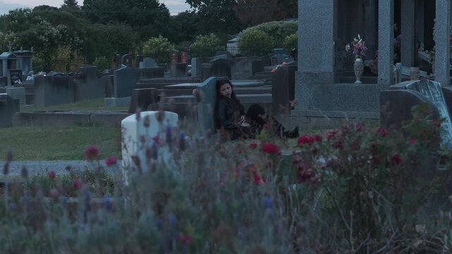 A girl sits beside a tombstone in a serene graveyard during a tranquil sunset. Flowers adorn the graves, creating a peaceful and reflective atmosphere. Soft, natural lighting enhances the mood.