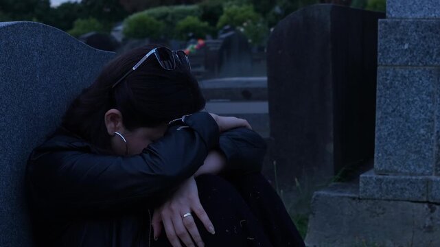 A solitary person in deep thought leans against a gravestone in a dimly lit graveyard. The scene captures a poignant, reflective mood, with cool lighting enhancing the somber setting.