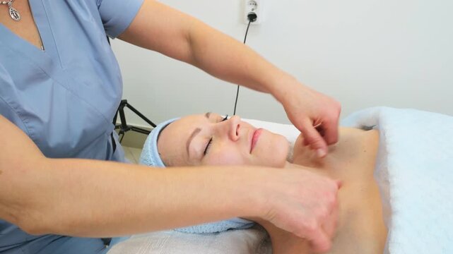 young woman lies on a spa bed enjoying a relaxing facial massage. A cosmetologist massages a female client's face for lymphatic drainage and lifting effects. Beauty, skincare concept.