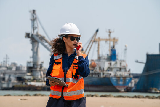 Woman wears white hard hat and orange safety vest. She holds tablet and speaks on mobile phone. woman engineer communicating via radio while inspecting maritime logistics at an industrial harbor.