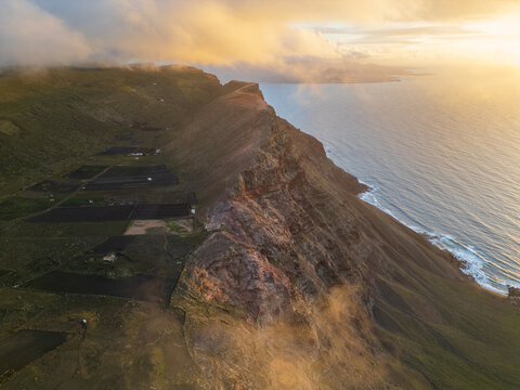 Aerial view of the dramatic Famara cliffs with golden sunset light hitting the volcanic rock face and clouds rolling over the ridge Lanzarote, Canarias, Spain.