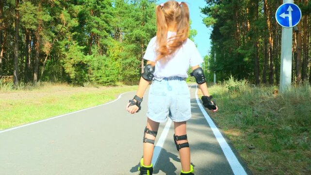 Child girl in casual clothes roller skating on road in forest on sunny summer day.