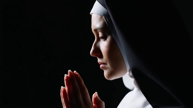 Slow motion close up profile of nun praying with hands clasped in dark studio interior with dramatic side lighting