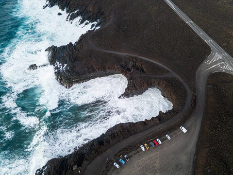 Aerial view of the rugged volcanic coastline with crashing white waves against dark basalt rocks and a coastal road in Lanzarote, Canarias, Spain.