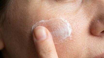 Fototapeta premium Detail of woman hand applying nourishing skincare cosmetic cream on face, facial skin texture close up detail.