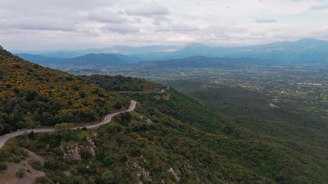 A cinematic highangle shot of a serpentine road curving through lush green mountains overlooking a vast valley under a cloudy sky in Baunei, Italy.