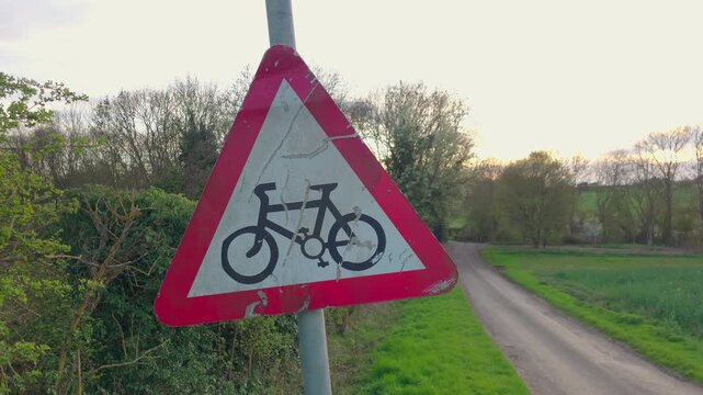 Cyclist warning road sign near narrow rural lane with countryside landscape and green fields at sunset. Traffic safety sign with bicycle symbol placed beside quiet country road with trees and hedges