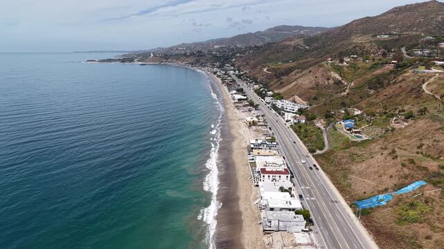 Cinematic 4K 10-bit aerial drone footage captured above the shoreline near Pacific Coast Highway in Malibu, featuring a smooth reverse dolly movement.