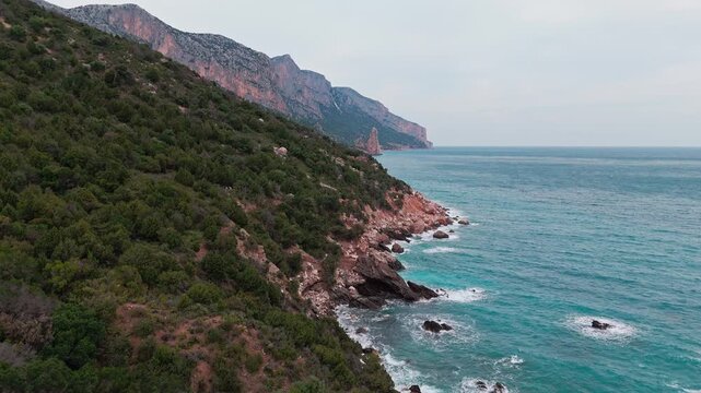 Stunning drone shot of the rocky coastline and limestone cliffs at Pedra Longa, Baunei, featuring turquoise Mediterranean waters and lush green vegetation.