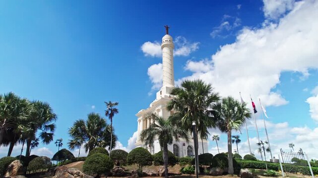 Low angle tracking shot through palm trees revealing the Monumento a los Heroes de la Restauraci&oacute;n under a blue sky with clouds in Santiago, Dominican Republic