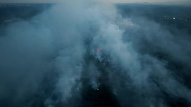 Smoke rises thick and grey above dark earth. Flames flicker beneath the haze, distant and fierce