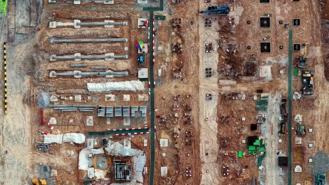 Aerial view of active construction site. Workers move among heavy machinery. Earthworks and concrete structures form orderly layout