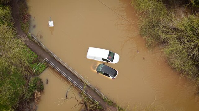 Storm flooding in UK submerging rural road and vehicles in muddy river water. Rising water levels trapping cars near pedestrian bridge during extreme rainfall. Climate change driving severe weather