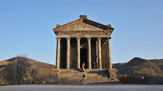 Ancient Garni temple with tall columns stands against clear blue sky. Person walks near the temple's entrance