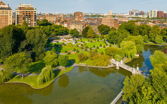 Aerial view of Public Garden with the Lagoon Bridge, people walking on paths, lush green trees, and the city skyline in Boston, Massachusetts, United States.