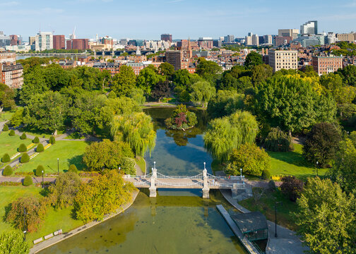 Aerial view of the Public Garden featuring the Lagoon Bridge over the pond with people walking along paths surrounded by trees and the city skyline in Boston, Massachusetts, United States.