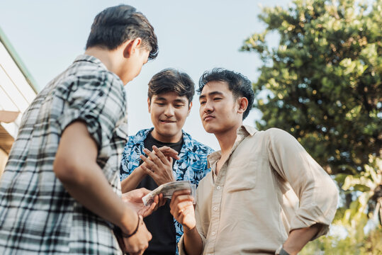 A hesitant young Asian man handing money to two intimidating men during a tense outdoor confrontation. Outdoor shot.