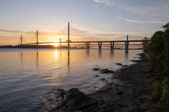 Aerial view of Forth Bridges spanning the Firth of Forth during a golden sunset with calm water and rocky shoreline West Lothian, UK.
