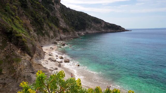 Aerial couple walking along pristine Filikuri beach with blooming flowers. Clear turquoise Ionian sea meets steep rocky coastal cliffs. Scenic Albanian Riviera travel landscape in Himar&euml;, Albania.