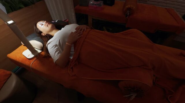 Woman receiving beauty treatment at a spa salon, comfortably lying on an orange massage bed with eye pads, enjoying relaxation and pampering during the cosmetic procedure