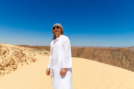 Arab man in national dress stands in the Sinai or Sahara desert and looks at the blue sky. Man in traditional thobe tobe thawb dishdasha kandura and sunglasses standing proudly in desert.